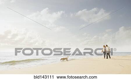 Stock Image - Caucasian couple walking dog on beach. Fotosearch