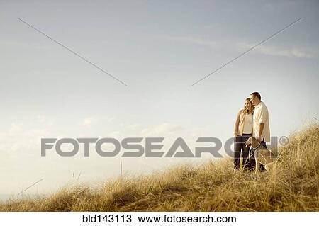 Stock Image - Caucasian couple walking dog on grassy dunes. Fotosearch