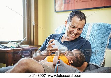 Stock Photo - Caucasian father feeding baby boy in living room. Fotosearch