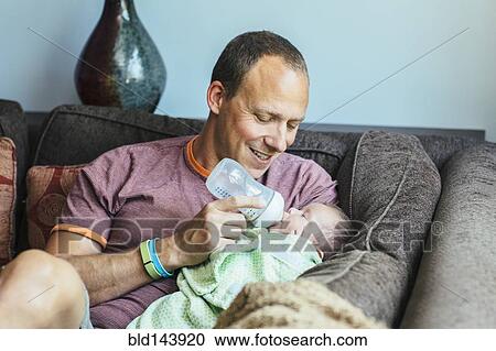 Stock Image - Caucasian father feeding baby boy on sofa. Fotosearch