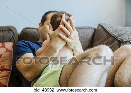 Stock Image - Caucasian father holding baby boy on sofa. Fotosearch