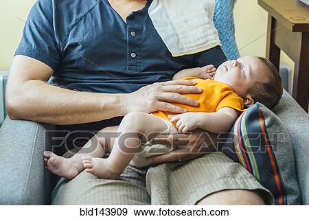 Stock Photo - Caucasian father holding sleeping baby boy in armchair. Fotosearch