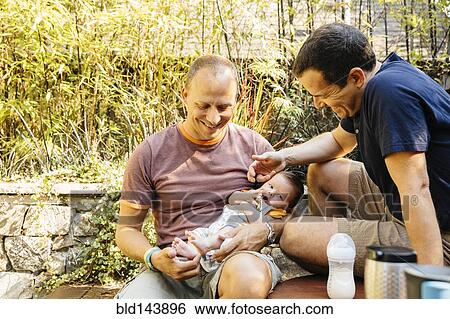 Stock Photograph - Caucasian gay couple admiring baby boy in backyard. Fotosearch