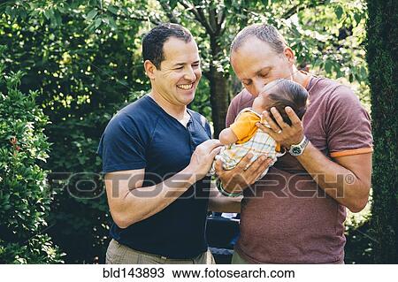 Stock Image - Caucasian gay couple kissing baby boy outdoors. Fotosearch