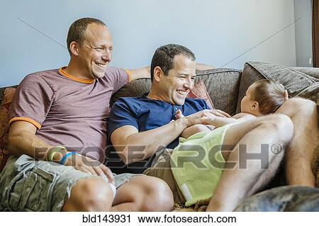 Stock Image - Caucasian gay couple playing with baby boy on sofa. Fotosearch