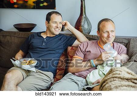 Stock Image - Caucasian gay couple relaxing with baby boy on sofa. Fotosearch
