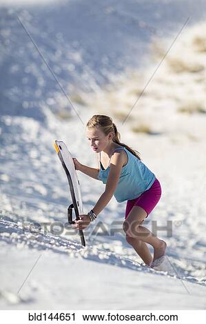 Stock Image - Caucasian girl climbing sand dune with sled. Fotosearch