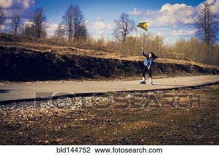 Stock Image - Caucasian girl on inline skates flying kite on rural road. Fotosearch