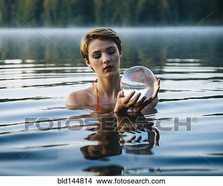 Picture - Caucasian woman holding crystal ball in still lake. Fotosearch