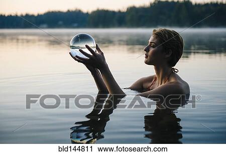 Stock Image - Caucasian woman holding crystal ball in still lake. Fotosearch