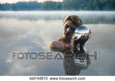 Stock Image - Caucasian woman holding crystal ball in still lake. Fotosearch