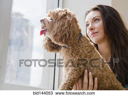 Stock Image - Caucasian woman looking out window with pet dog. Fotosearch