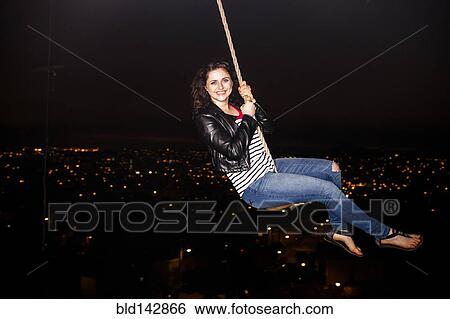 Stock Photograph - Caucasian woman on rope swinging over scenic view of cityscape. Fotosearch