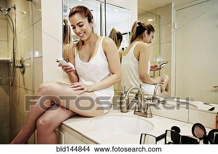 Picture - Caucasian woman using cell phone on bathroom counter. Fotosearch