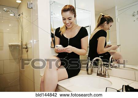 Stock Image - Caucasian woman using cell phone on bathroom counter. Fotosearch
