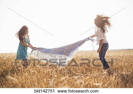 Stock Image - Caucasian women spreading blanket in rural field. Fotosearch