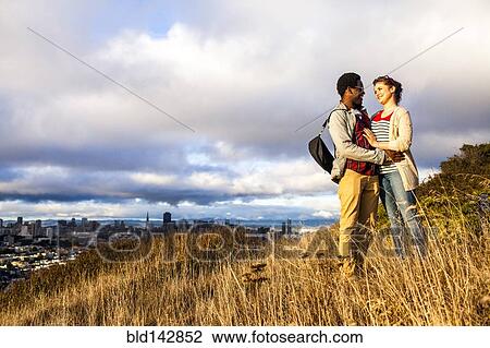Stock Image - Couple hugging on grassy hill overlooking cityscape. Fotosearch