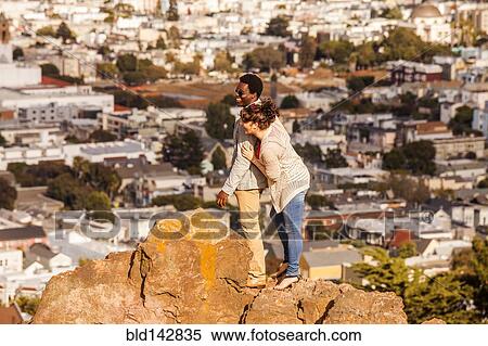 Stock Photography - Couple hugging on rock overlooking scenic view of cityscape. Fotosearch