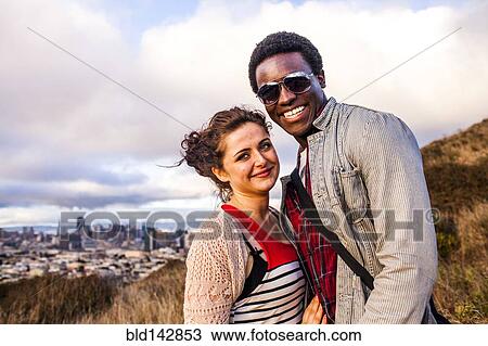 Stock Image - Couple smiling on grassy hill overlooking cityscape. Fotosearch