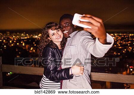 Stock Photo - Couple taking cell phone selfie near scenic view of cityscape at night. Fotosearch