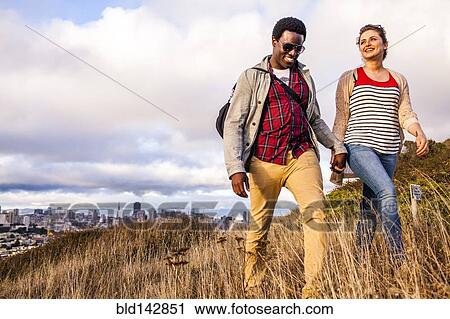 Stock Image - Couple walking on grassy hill overlooking cityscape. Fotosearch