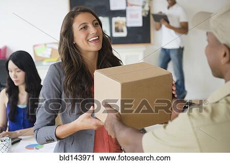 Stock Image - Delivery man handing cardboard box to businesswoman in office. Fotosearch