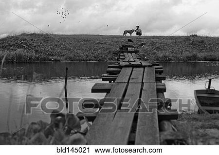 Stock Image - Girl petting dog near rustic wooden bridge crossing river. Fotosearch