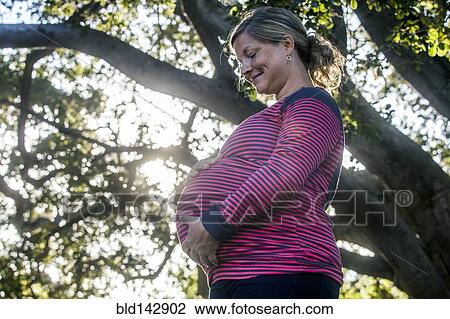 Stock Image - Low angle view of pregnant Caucasian mother holding her stomach outdoors. Fotosearch