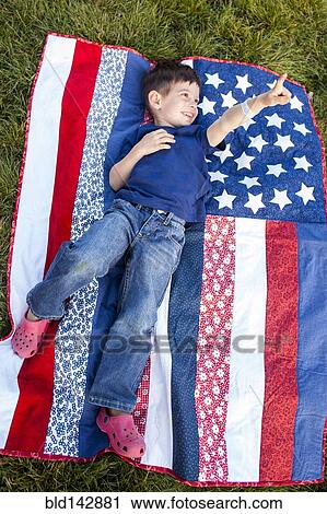 Stock Image - Mixed race boy pointing up on American flag blanket on grass. Fotosearch