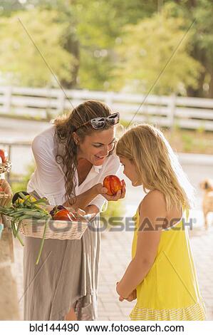 Stock Image - Mixed race mother and daughter smelling produce at farmers market. Fotosearch