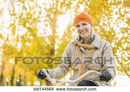 Picture - Older Caucasian woman riding bicycle under autumn trees. Fotosearch
