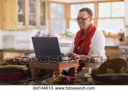 Stock Image - Older mixed race woman using laptop in home office. Fotosearch