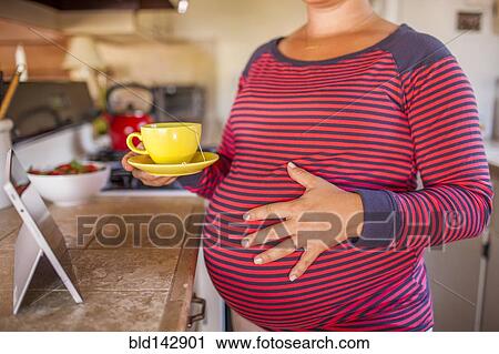 Stock Image - Pregnant Caucasian mother holding cup of tea in kitchen. Fotosearch