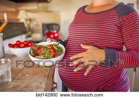 Stock Image - Pregnant Caucasian mother holding salad in kitchen. Fotosearch