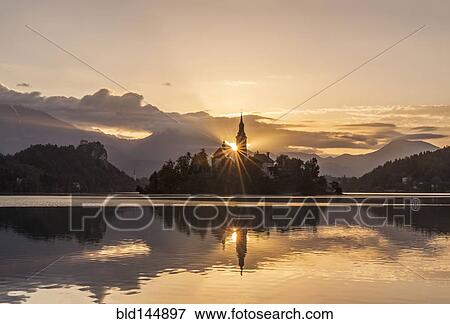 Stock Photo - Sunrise over church tower reflected in still lake, Bled, Upper Carniola, Slovenia. Fotosearch