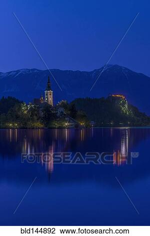 Stock Image - Village church and buildings reflected in still lake at night, Bled, Upper Carniola, Slovenia. Fotosearch
