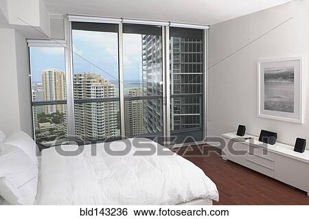 Stock Photograph - Windows overlooking urban high rise buildings in modern bedroom. Fotosearch