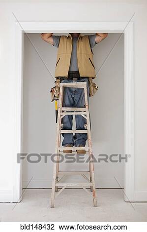 Stock Image - Caucasian carpenter standing on ladder in doorway. Fotosearch