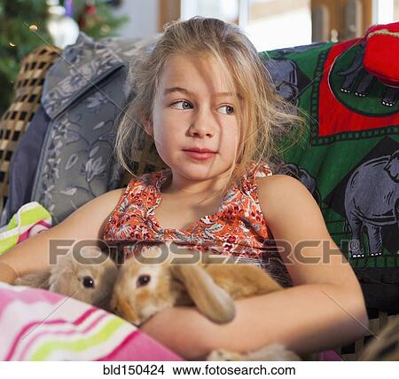 Picture - Caucasian girl holding pet rabbits on sofa. Fotosearch