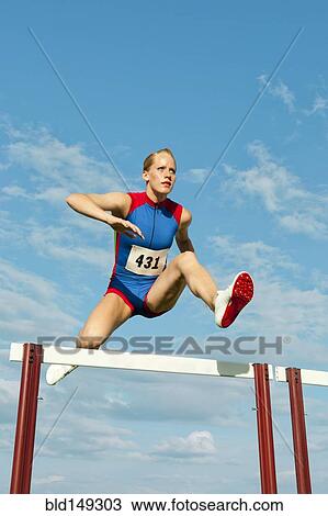 Stock Image - Caucasian runner jumping over hurdles on track. Fotosearch