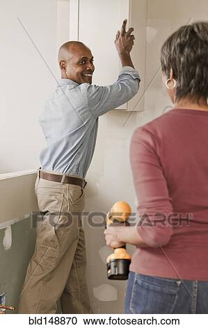 Stock Image - Couple hanging cabinets in unfinished room. Fotosearch