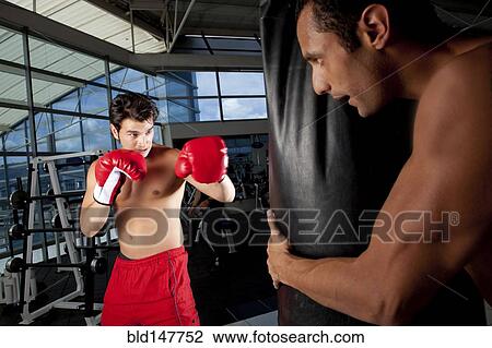 Stock Image - Hispanic man using punching bag in health club. Fotosearch