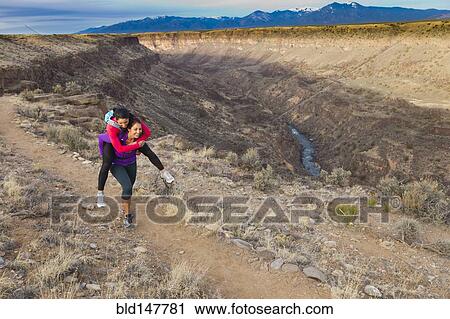 Stock Image - Hispanic woman carrying friend on her back in remote area. Fotosearch