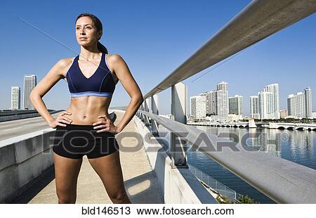 Stock Image - Hispanic woman in sportswear standing near urban waterfront. Fotosearch