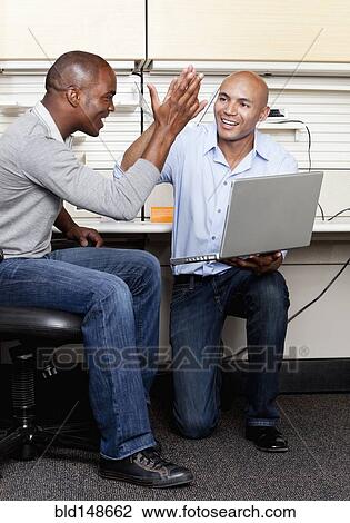 Stock Image - Mixed race businessmen high fiving in office cubicle. Fotosearch