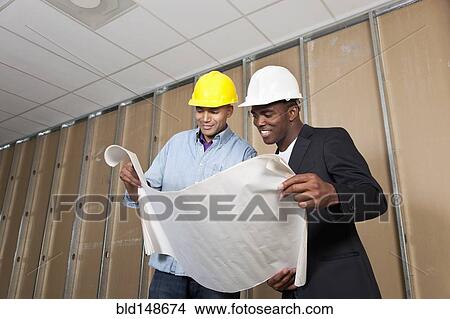 Mixed race businessmen looking at blueprints in room under construction View Large Photo Image Picture - Mixed race businessmen looking at blueprints in room under construction. Fotosearch