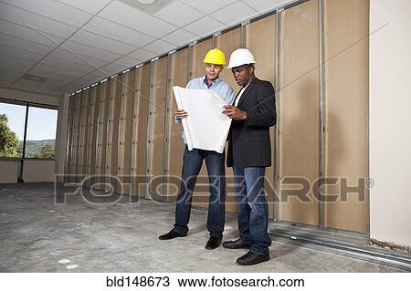 Stock Image - Mixed race businessmen looking at blueprints in room under construction. Fotosearch