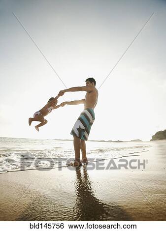 Stock Photograph - Mixed race father swinging daughter on beach. Fotosearch