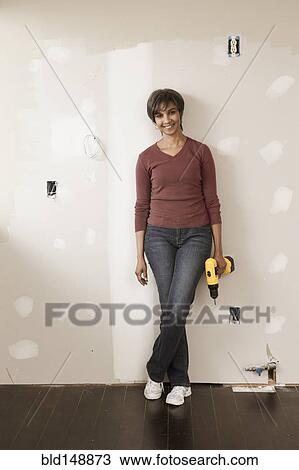 Stock Image - Mixed race woman holding cordless drill in unfinished room. Fotosearch