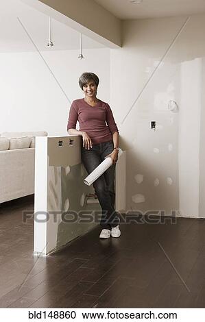 Stock Image - Mixed race woman standing in unfinished room with blueprints. Fotosearch